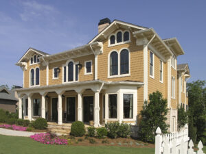 Two-story home with tan lap siding, multiple styles of windows including arched and a bay window, with windows and front porch columns painted white
