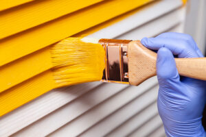 Painting Company Dallas-Fort Worth TX Closeup female hand in purple rubber glove with paintbrush painting natural wooden door with yellow paint.