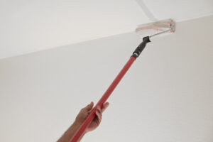 Hand of a man holding a paint roller on a long telescopic pole to paint the ceiling